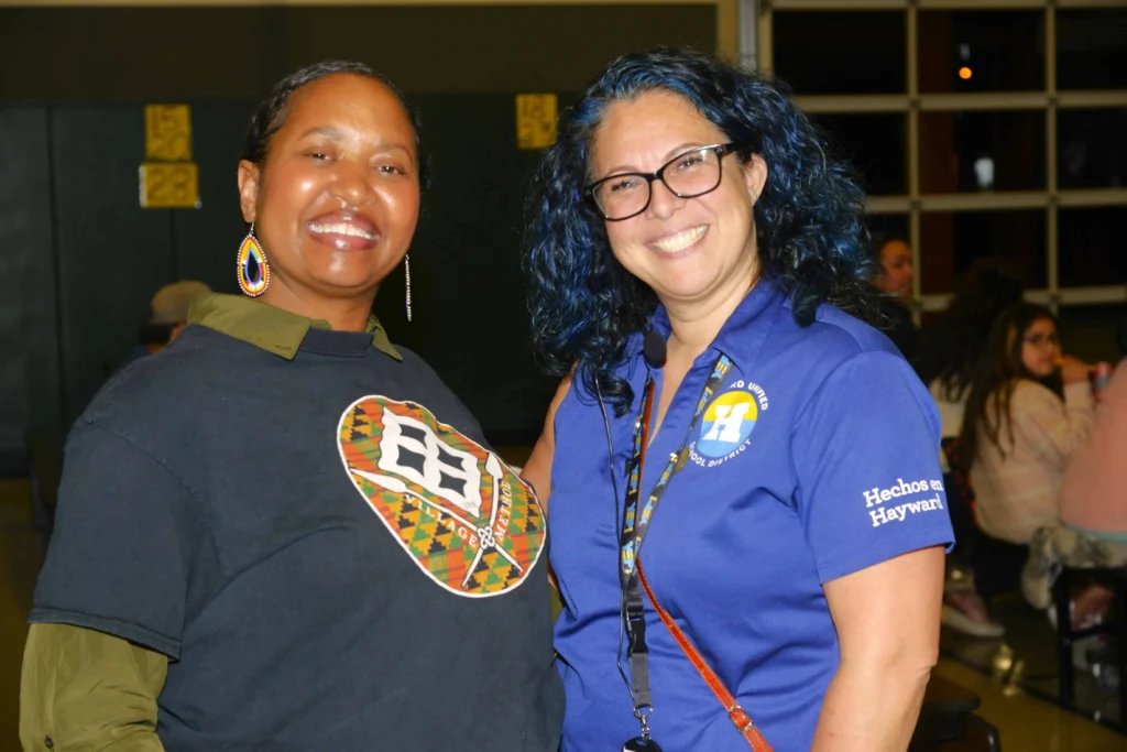 Two women smiling at the camera. One is wearing a black T-shirt with a colorful logo, and the other is wearing a blue polo shirt with "Hechos en Hayward" embroidered on the sleeve, standing in a gymnasium.