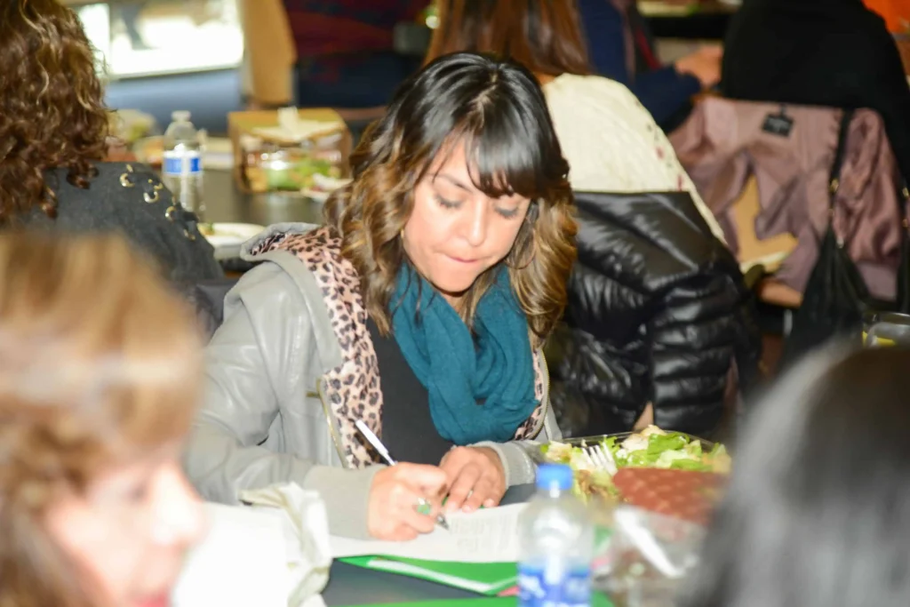 A woman in a teal scarf writing notes while sitting at a conference or workshop.