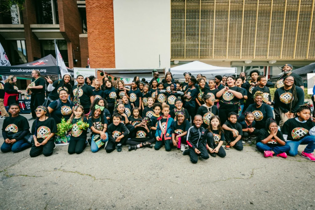 A large group of children and adults gathers outside, many wearing matching T-shirts, creating a lively and spirited group portrait.