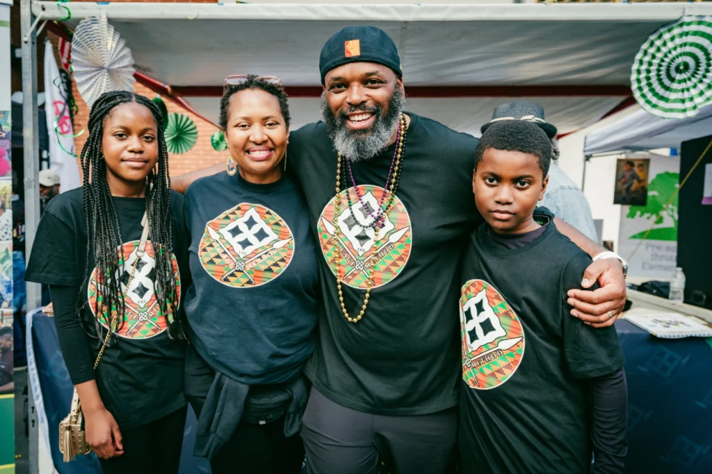 A family or small group poses warmly at an outdoor event, all wearing matching culturally themed shirts, highlighting unity and pride.