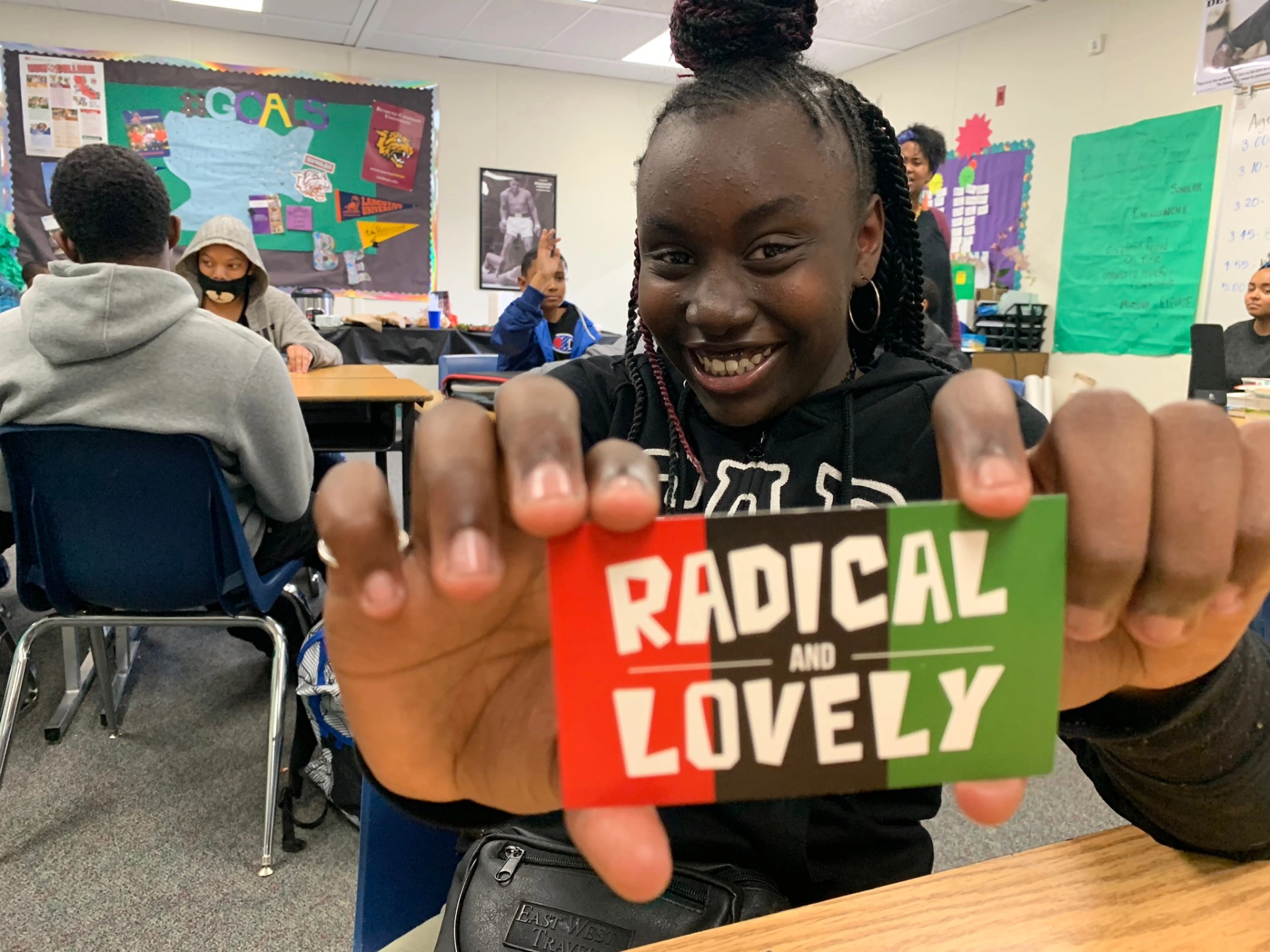 A young girl sitting in a classroom holding a card that says "Radical and Lovely" with a big smile on her face.