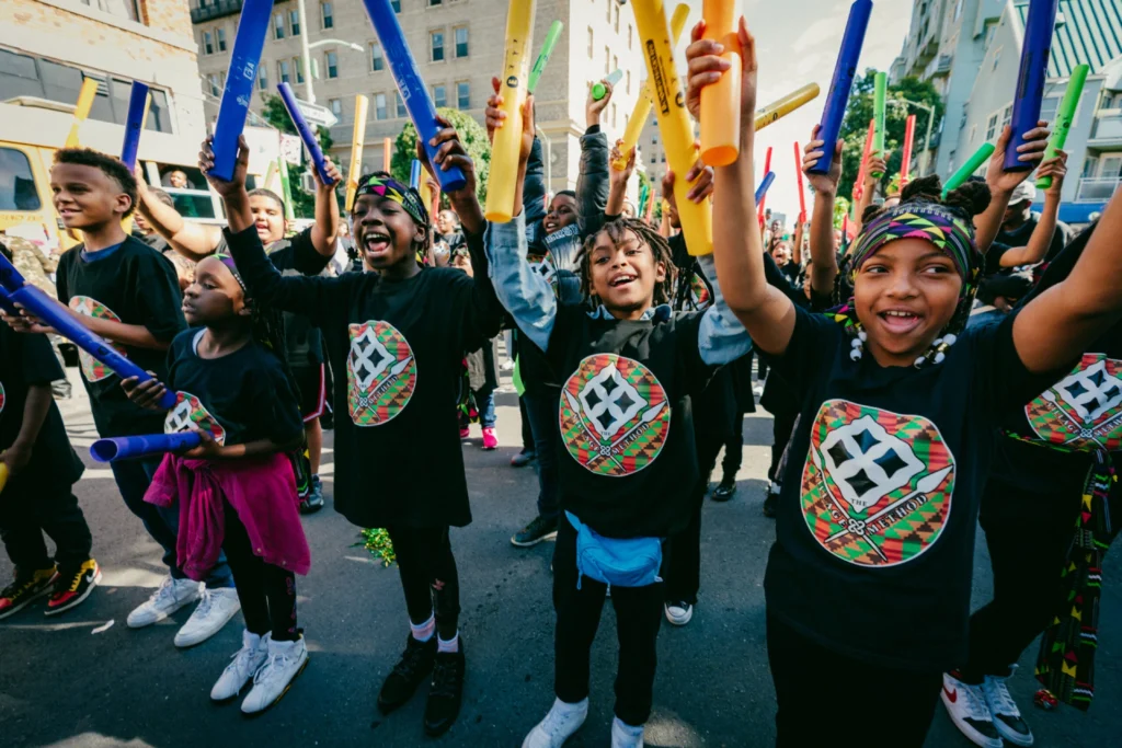 A group of children joyfully participates in an outdoor event, raising colorful sticks in the air and wearing matching T-shirts, radiating energy and excitement.
