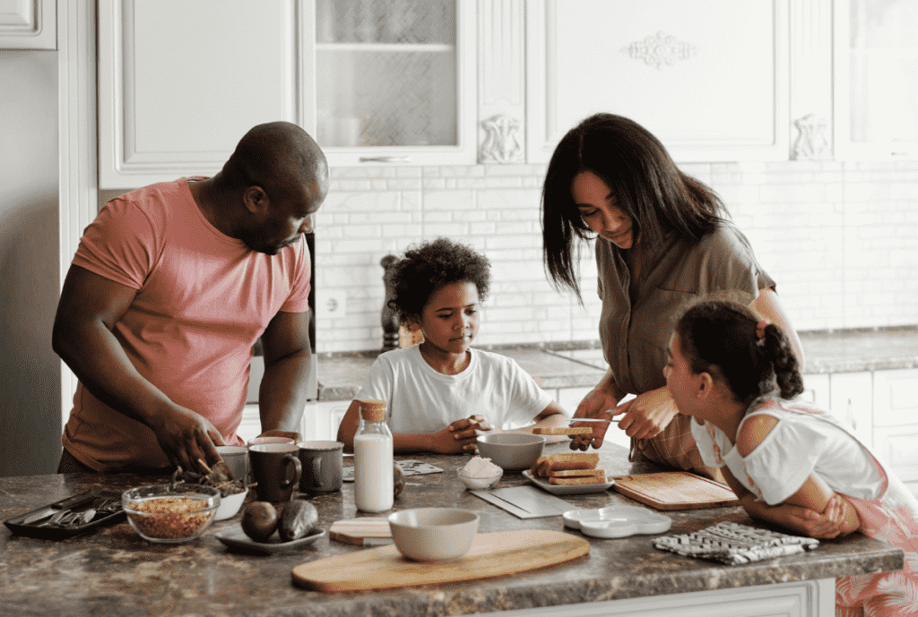 A Black family of four—a father, mother, and two children—gathering around a kitchen island to prepare a healthy breakfast together in a bright, modern white kitchen in this post from The Village Method about building routines, consistency and connection at home.