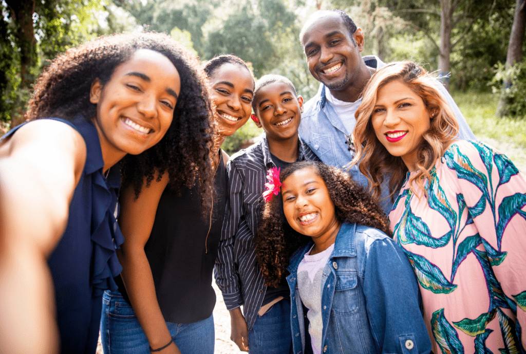 A multi-generational Black and Latino family smiling together for a group selfie outdoors in a sunny park, representing a strong community and supportive virtual village.