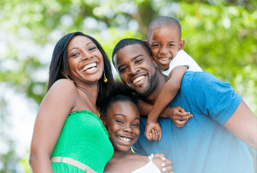 A joyful Black family of four—a mother, father, daughter, and young son—smiling and hugging outdoors against a blurred green leafy background, representing a supportive home environment in a post on The Village Method about Mid-Year School Check-In for Families