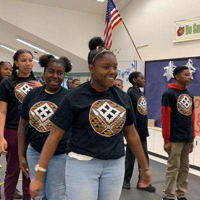 Students walking in a line on stage wearing matching black shirts with colorful logos.