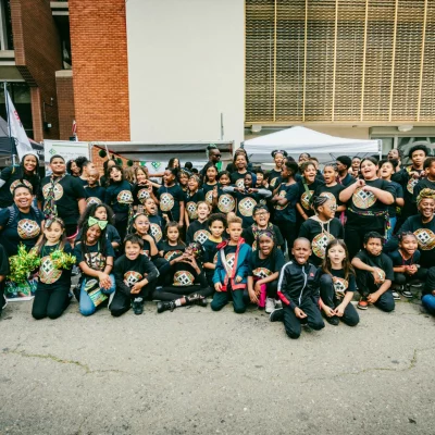 A large group of children and adults gathers outside, many wearing matching T-shirts, creating a lively and spirited group portrait.