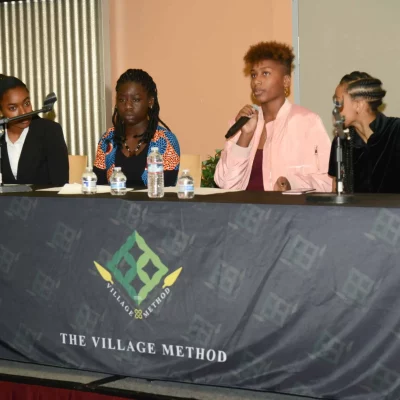 A panel of young women speaking at an event, with The Village Method logo displayed on the table.